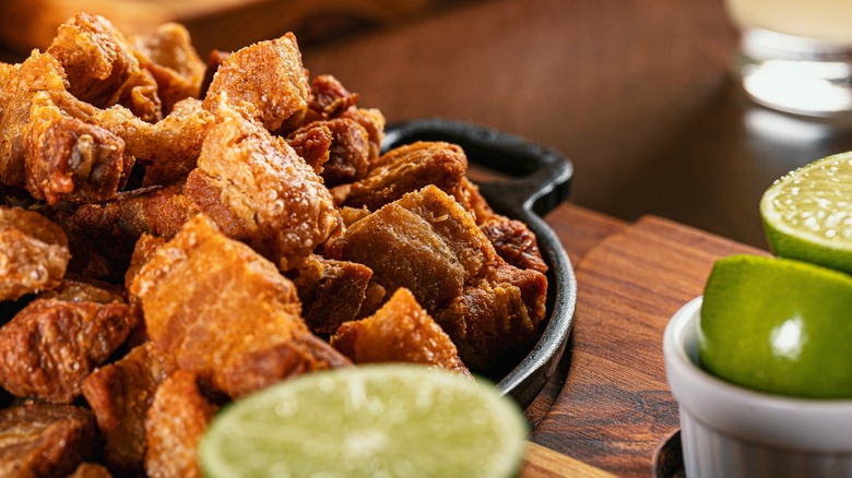 A cast iron dish of super crispy, golden brown pork cracklings, surrounded by sliced limes.