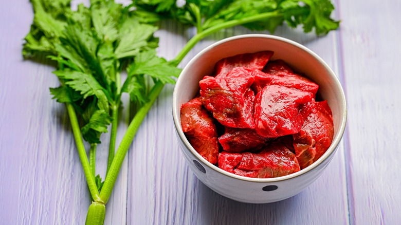 Raw chunks of beef in ceramic bowl next to celery on wooden table.