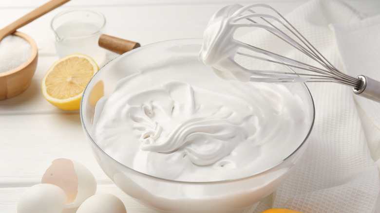 Glass bowl of whipped cream with whisk on white table with other ingredients