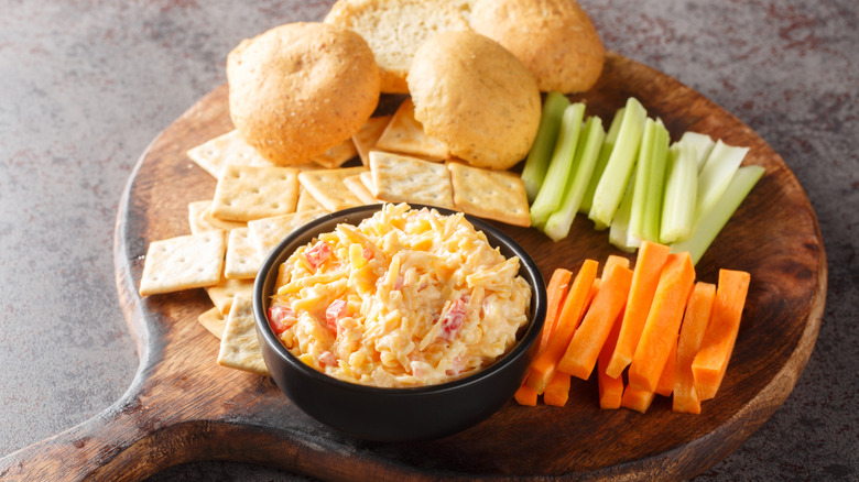 Pimento cheese dip alongside celery, carrot sticks, and crackers on a cutting board.