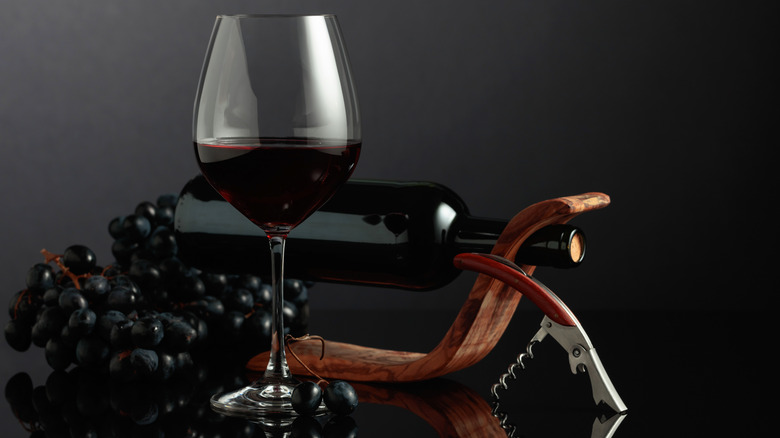 Glass of red wine in front of decorative wine bottle holder, with grapes and wine key in background.