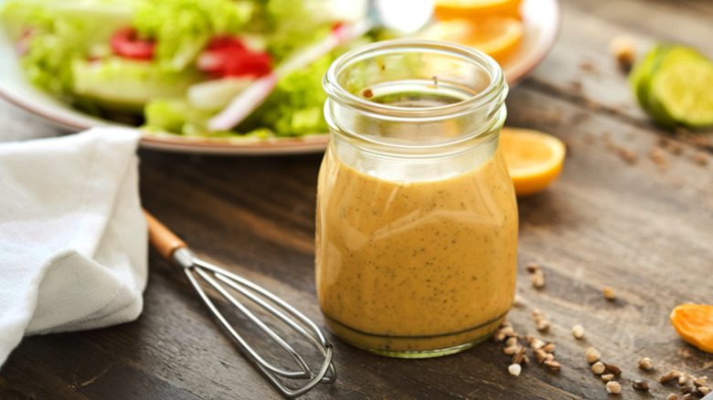 A jar of honey mustard on a table with a salad and whisk