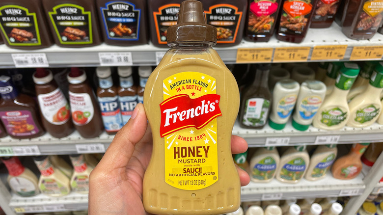 Close up of hand holding a bottle of French's honey mustard in front of a condiment aisle