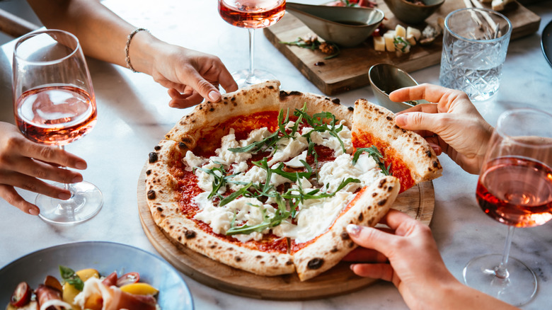 Marble countertop with glasses of wine, a pizza, and hands reaching for slices