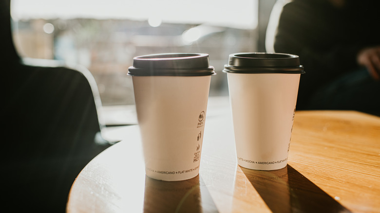 Two cups of coffee in to-go cups sit on top of a wooden table.