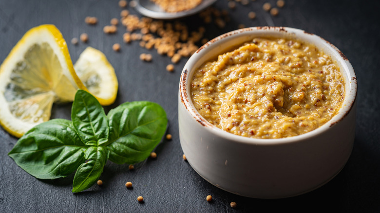A white ramekin of grainy mustard; a lemon twist and herb leaves are also styled in the frame.
