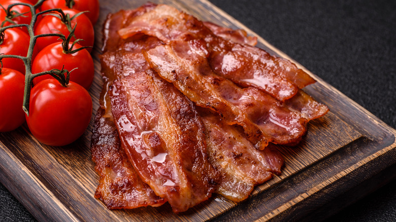 A few slices of cooked bacon on a wood bard with tomatoes on the vine next to them.
