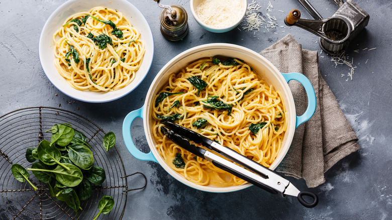 Tongs resting in a pot of spinach pasta next to a bowl of pasta, a napkin, and cheese