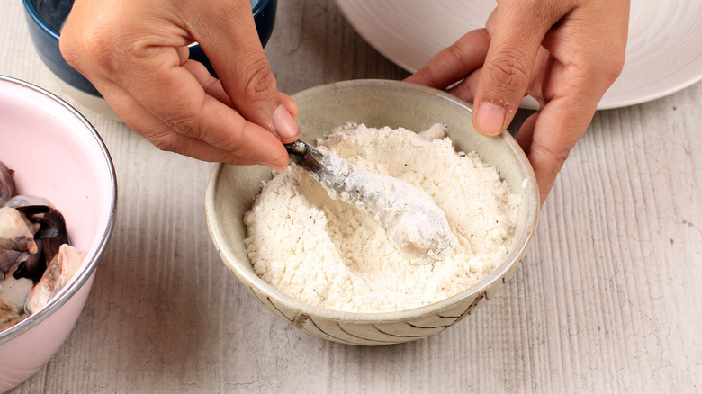 Hands breading a piece of shrimp in a bowl