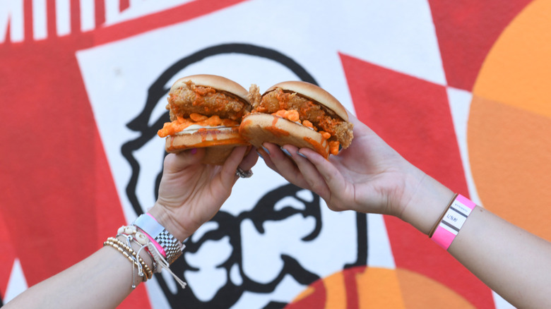 Two people each hold the KFC Cheetos Sandwich up in front of a KFC sign