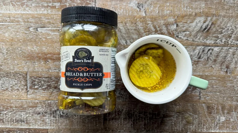 A jar of Boar's Head bread and butter pickles next to a cup holding pickles on a table
