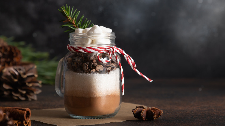 Small glass mason jar filled with layered ingredients of hot cocoa with pine cones in background