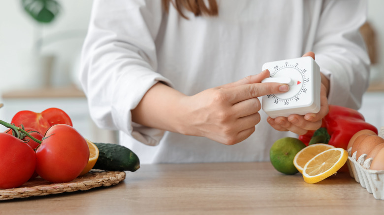 A person's hands are seen setting a kitchen timer to 15 minutes over a counter topped with eggs, tomatoes, and other ingredients.