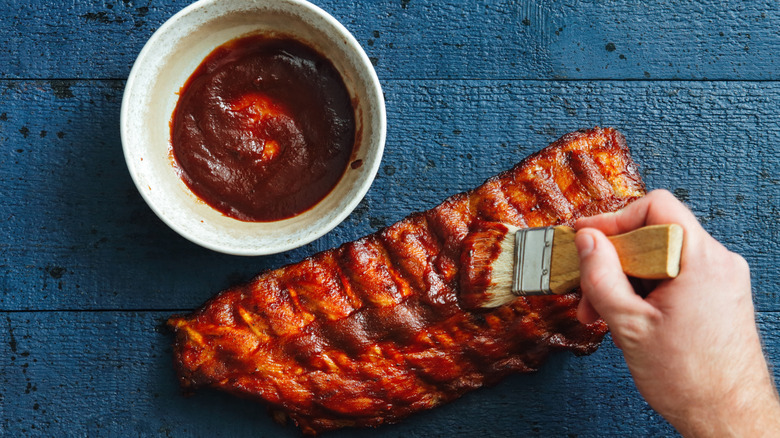Hand holding brush slathering BBQ pork ribs with sauce next to small bowl of BBQ sauce