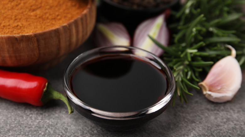 A close-up of a bowl of soy sauce and three garlic cloves, surrounded by a sprig of rosemary, a fresh chile, and a bowl of cayenne pepper