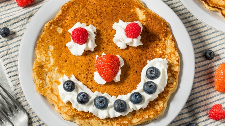A breakfast pancake for kids on a plate with berries and cream arranged into a smiley face.