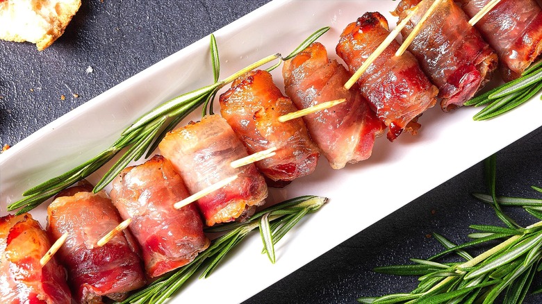 Bacon wrapped appetizers on a long white tray with sprigs of fresh rosemary