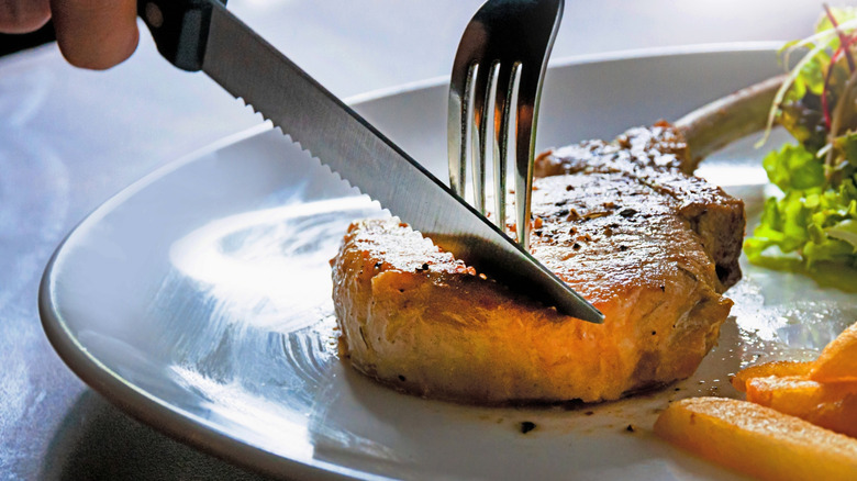 A knife and fork cut into a pork chop on a plate.