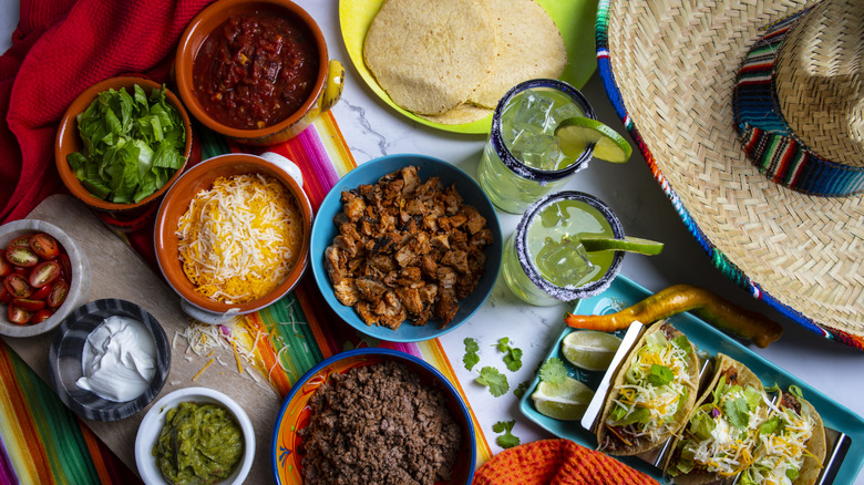 Serving of three tacos along with bowls of additional ingredients and tortillas to make your own plus two margaritas