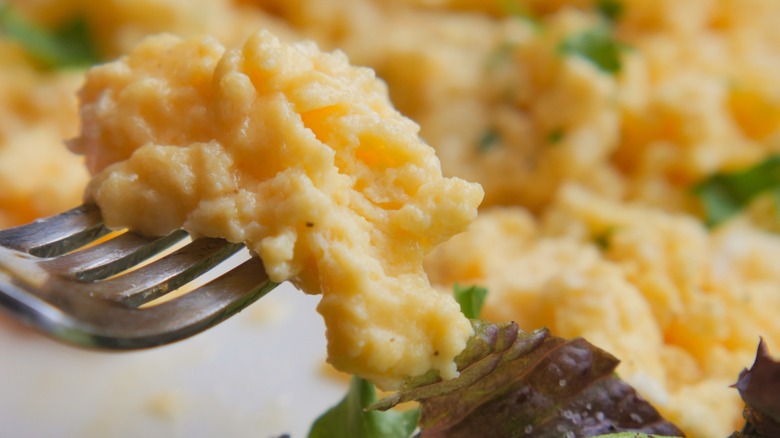 Golden yellow scrambled eggs on a fork with vegetables and more eggs behind it.