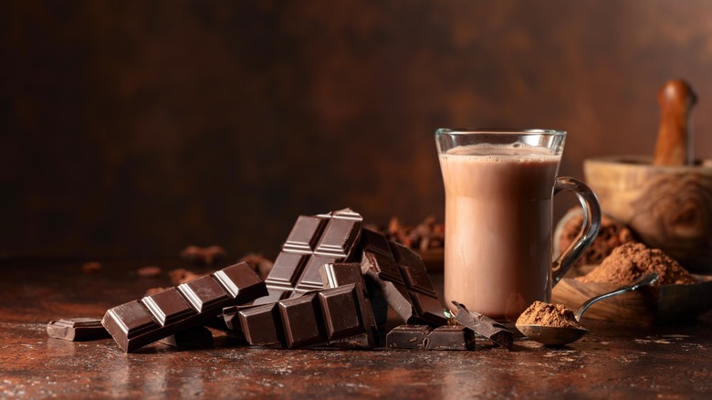 A clear, glass mug of hot chocolate with chocolate squares and cocoa powder in the frame.