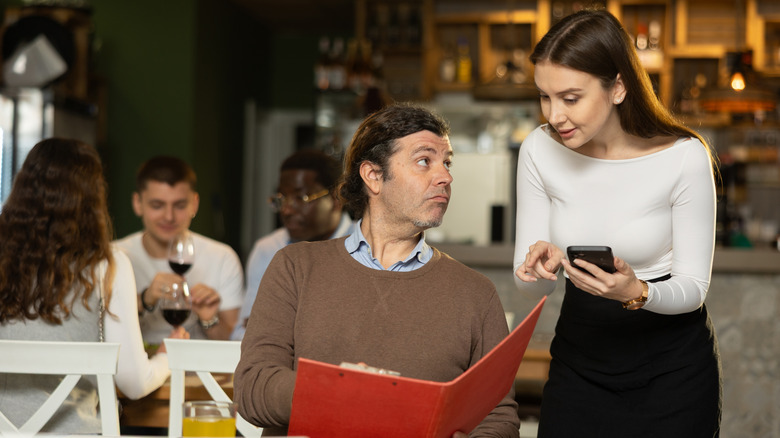 Person in restaurant points at menu and looks at waitstaff as they answer question