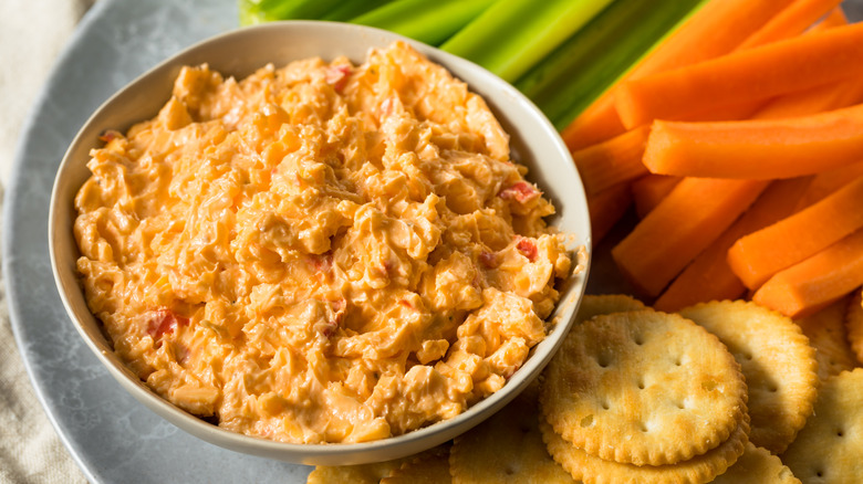 Pimento cheese in a bowl with Ritz crackers and carrot and celery sticks.