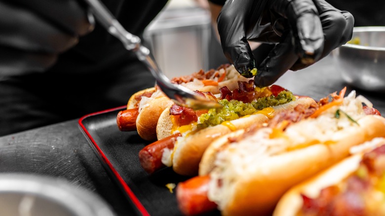 Chef wearing black gloves adding toppings onto various plated hot dogs