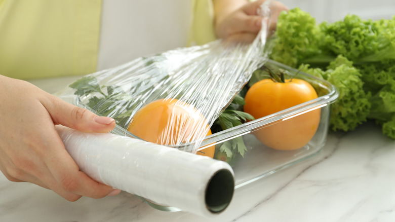 A person's hands are seen covering a glass dish of vegetables with cling wrap.