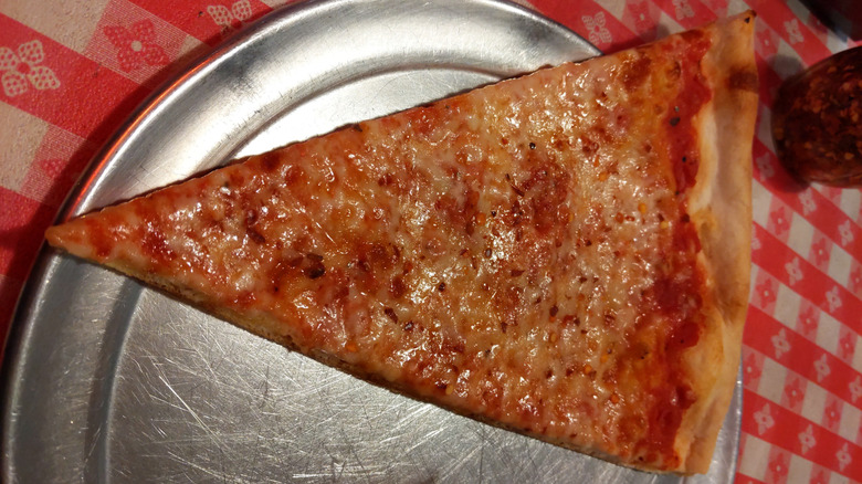 A cheese pizza slice from Carmine's in Dallas, Texas, sits on a metal plate on a red-and-white checkerboard tablecloth.