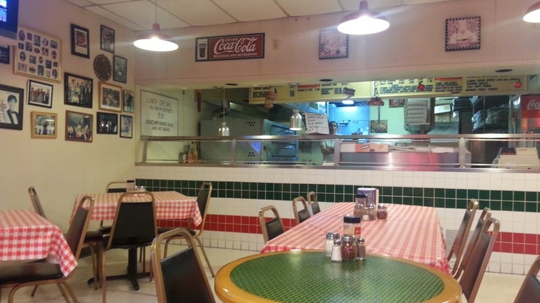 The inside of Carmine's Pizzeria in North Dallas with red-and-white checkered tablecloths, framed pictures hanging on the wall, and a large analog menu board behind the counter. A serious-looking chef stands next to the pizza ovens, wearing a white apron and shirt.