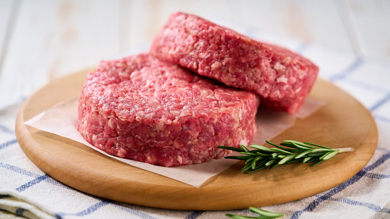 Uncooked burger patties are unevenly stacked on a sheet of parchment paper on a round wooden board