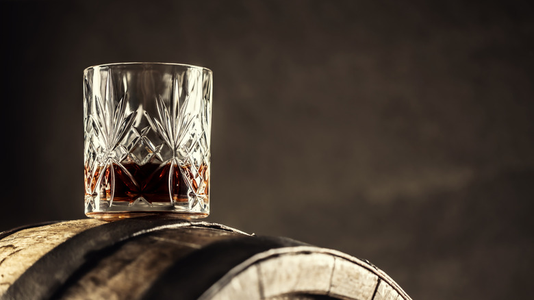 Tumbler glass of dark whiskey on top of an oak barrel