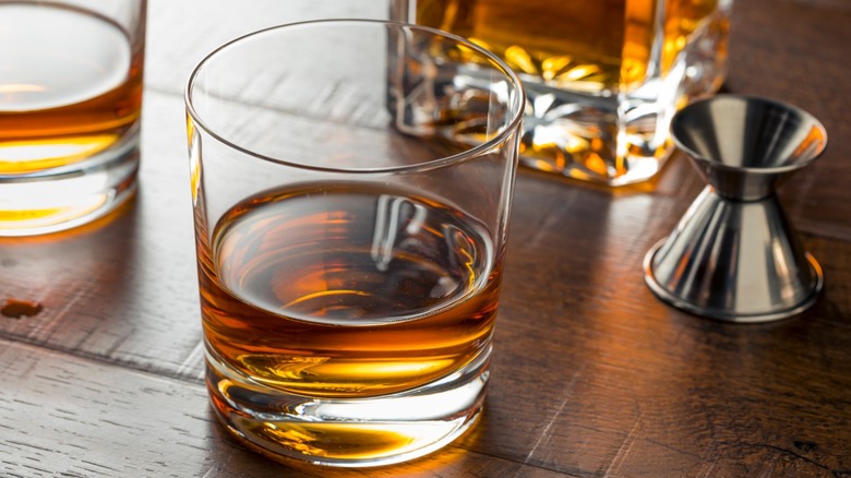 Pours of whiskey neat in rocks glasses sitting on a wood counter with a bottle of whiskey and a jigger.