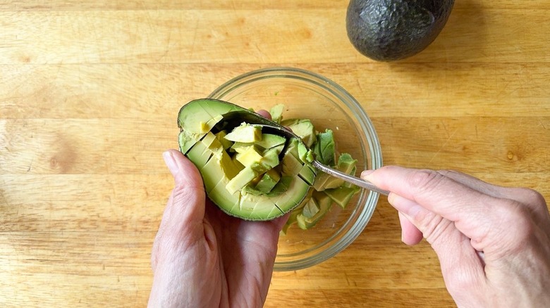 Scooping avocado cubes out of shell into glass bowl with spoon