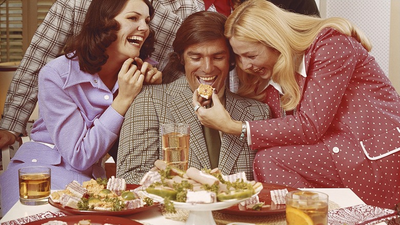 Vintage picture of three people laughing, with one woman feeding a man a piece of food