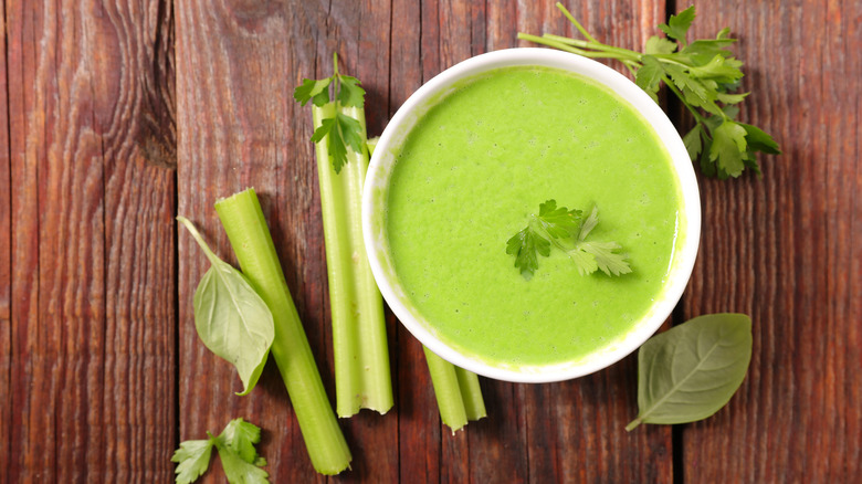 A bowl of green soup surrounded by stalks of celery and various herbs