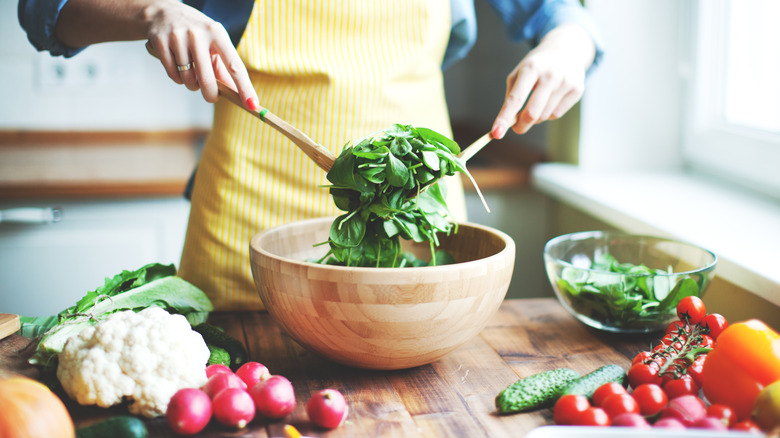 An apron-wearing person's hands are seen tossing salad greens in a wooden bowl; other veggies are arranged nearby.