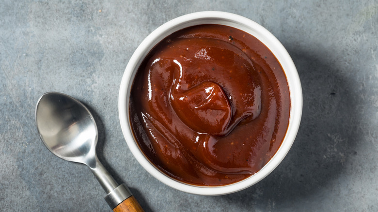 Barbecue sauce in a white ramekin next to a spoon on a gray surface
