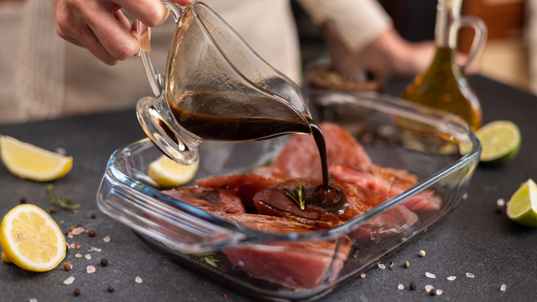 Chef pouring brown marinade onto meat in a glass dish on a slate countertop