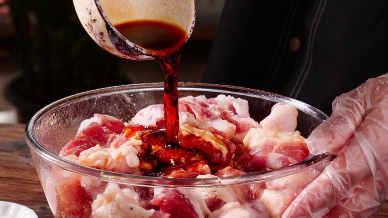 Person wearing gloves pouring marinade into a glass bowl of raw meat