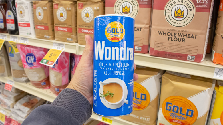 A hand holding up a blue canister of Gold Mill Wondra Quick-Mixing Flour in front of rows of other flour brands at the supermarket.