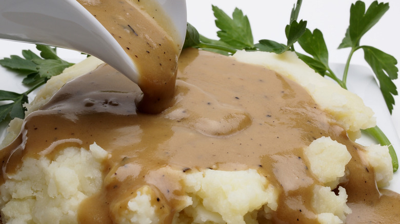 A close-up of a gravy boat pouring turkey gravy over a mound of mashed potatoes, a few sprigs of parsley in the background.