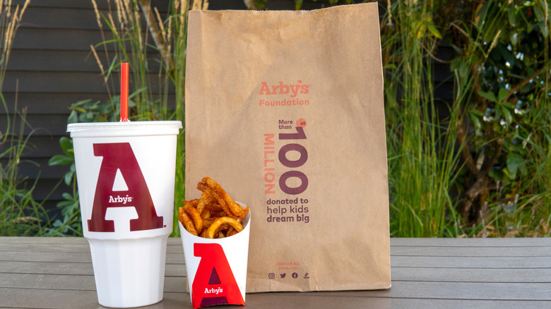 An Arby's takeout brown paper bag with drink and fries on a table outdoors