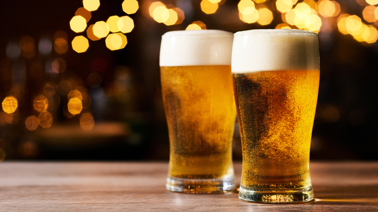 Two pints of beer siiting on a wooden table with lights in the background.