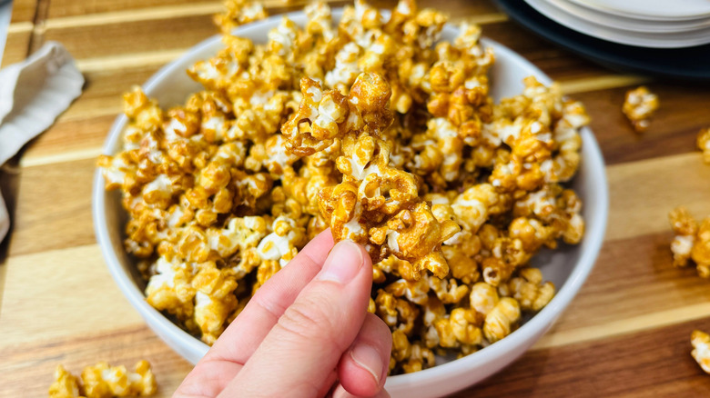 Hand holding caramel popcorn piece in front of bowl of caramel popcorn