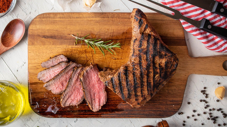 A sliced, charred tri-tip on a cutting board surrounded by peppercorns, rosemary, olive oil,