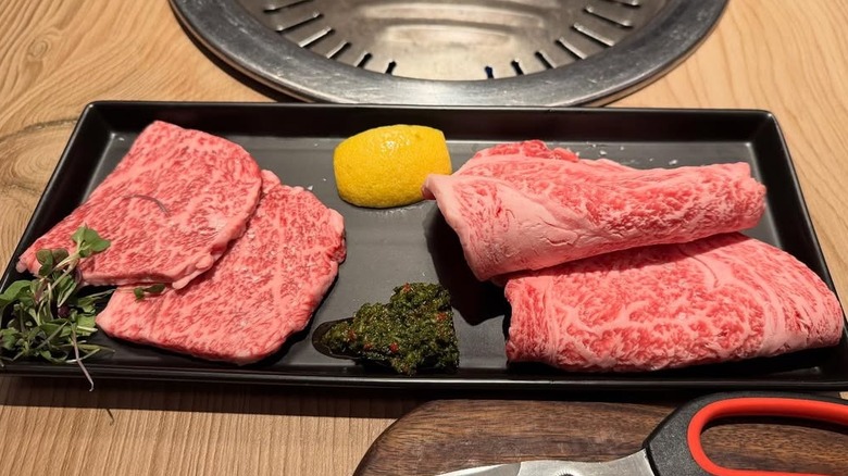 Sliced raw wagyu beef on black rectangular platter with lemon wedge and sauce in front of an on-table grill.