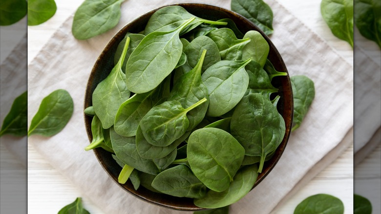 Raw spinach in a wooden bowl on a folded white tea towel with some leaves scattered on towel and table