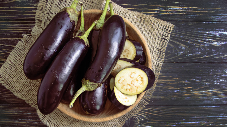 A wooden bowl containing several whole eggplants and several eggplant slices